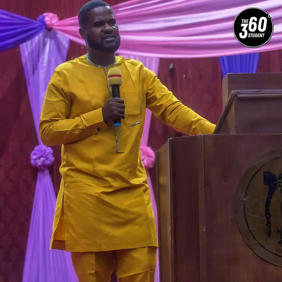 A speaker in gold traditional dress addressing the 360 Degree Student Conference from the podium.